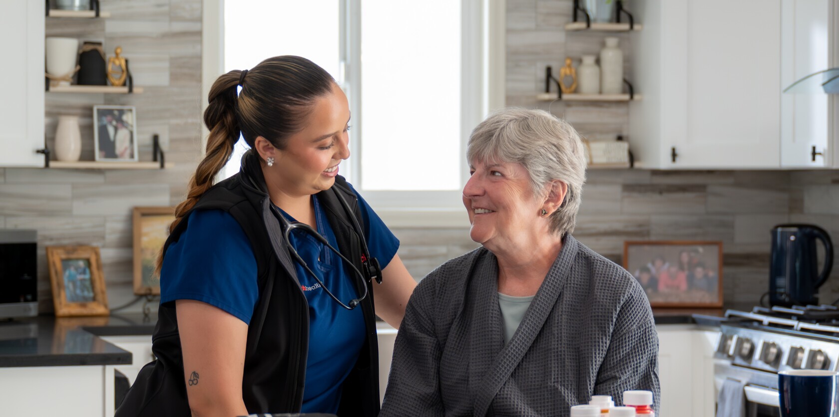 DispatchHealth provider in blue scrubs with stethoscope sitting with elderly female patient in modern kitchen, both smiling during in-home medical consultation
